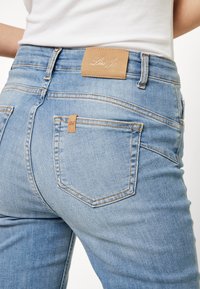 Jeans de mezclilla azul claro con una cintura alta, parche de cuero marrón con logo en la parte posterior y costuras en contraste en los bolsillos.