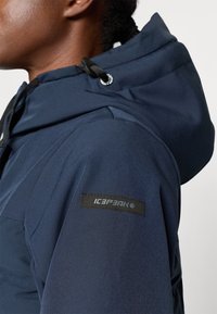Chaqueta azul marino con capucha, con una tela texturizada, cordón ajustable y una etiqueta de marca negra en el hombro.