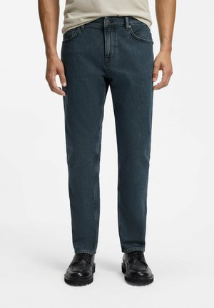 Mand iført mørkeblå jeans og sorte lædersko, stående mod en ensfarvet hvid baggrund.