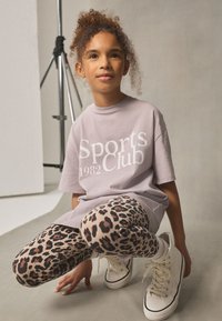 Light purple t-shirt with "Sports Club 1982" text; leopard print leggings; white sneakers. Model poses in a crouched position.