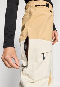 Beige en crême waterdichte broek met een zijrits, verstelbare tailleband en minimale hardware. Glad textuur en praktisch ontwerp van de zakken.