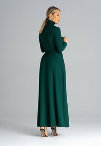 Długa, zielona sukienka z wysokim kołnierzem, długimi rękawami i ściągniętym w talii krojem. Materiał wydaje się gładki, a spódnica płynnie opada do podłogi.