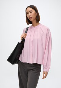 Rosa bluse med lange ermer og rund krage, med knappestolpe, kombinert med grå bukser og en svart håndveske. Glatt tekstur.