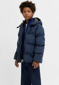 Tumši zils pūkains jaka ar kapuci, priekšā rāvējslēdzēja aizdare, divas sānu kabatas un kontrastējoša pelēka oderējuma iekšpuse. Teksturēta auduma.