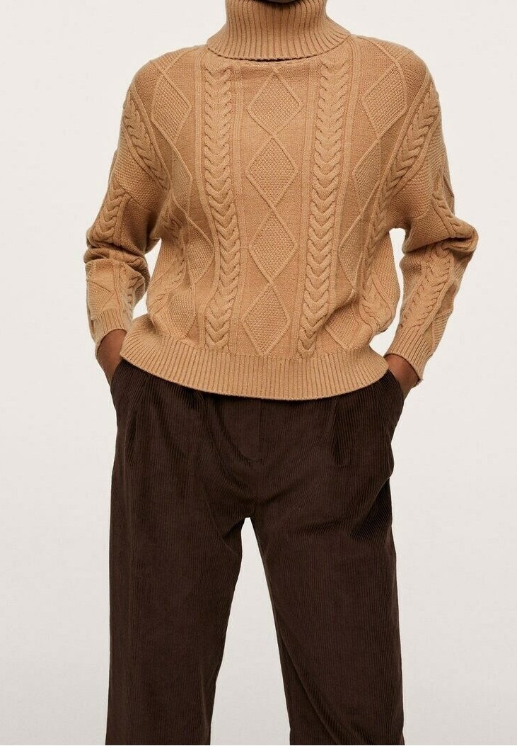 Beige gestrickter Pullover mit Stehkragen, der ein Rautenmuster und ein Zopfmuster verfügt, kombiniert mit dunkelbraunen Cordhosen.