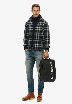 Black waterproof backpack with "SUPERDRY" logo, plaid flannel shirt in navy and green, dark jeans, and brown lace-up boots.