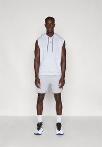 Sweatshirt cinzenta sem mangas com cordão e logótipo, combinada com calções cinzentos claros. Modelo usa ténis brancos com detalhes em preto.