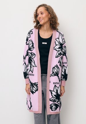 Vrouw glimlachend, draagt een lange roze cardigan met zwart-witte bloemmotieven over een zwarte top en grijze broek, staande tegen een witte achtergrond.