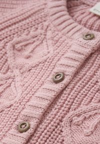 Ljusrosa stickad kofta med ribbat halslinning, texturerade mönster och träknappar för stängning. Mjuk, kraftig garn ger värme.