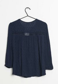 Granatowa bluzka z rękawami do połowy przedramienia, teksturowana tkanina z małymi kropkami oraz koronkowy panel na wysokości klatki piersiowej. Zawieszona na czarnym haczyku.