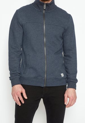 Homme portant une veste zippée bleu foncé avec des poches et un pantalon noir, debout devant un fond blanc uni.