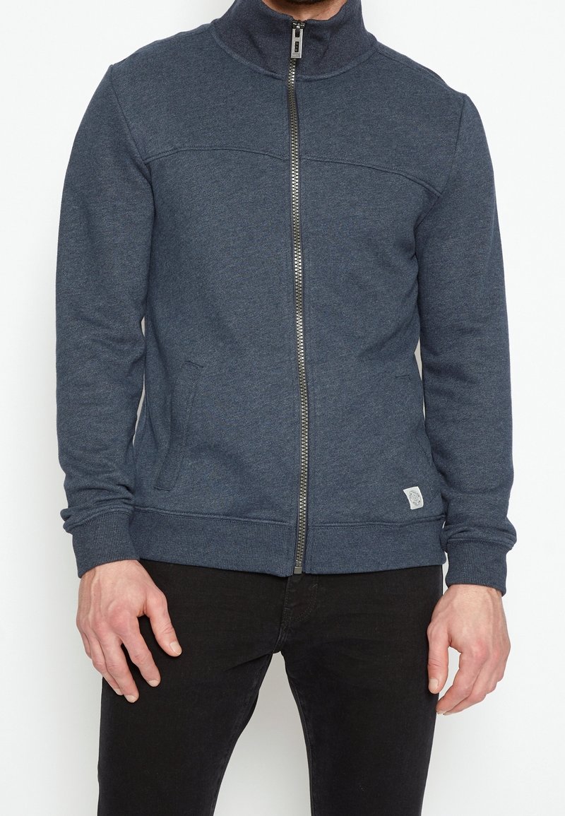 Homme portant une veste zippée bleu foncé avec des poches et un pantalon noir, debout devant un fond blanc uni.
