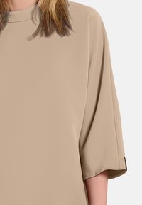 Beige ylhäällä oleva paita, jossa on korkea kaulus ja leveät, lyhyet hihat. Valmistettu sileästä kankaasta, jossa on hieno kiilto. Minimalistinen muotoilu ilman kuvioita.