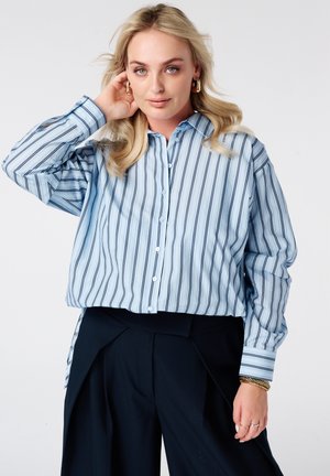 Femme blonde portant une chemise boutonnée à rayures bleu clair et un pantalon noir, avec des boucles d'oreilles et bracelets dorés, posant avec une main près du visage.
