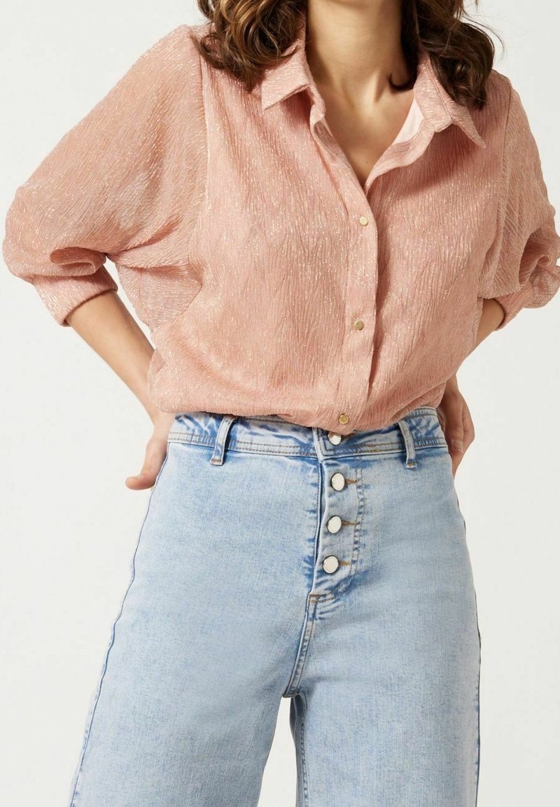 Femme portant un chemisier texturé rose clair à manches longues, rentré dans un jean en denim bleu clair taille haute avec quatre boutons blancs.