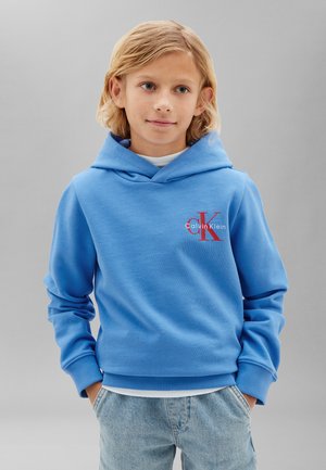 Blond dziecko w niebieskiej bluzie z kapturem Calvin Klein i jasnoniebieskich dżinsach, stojące z rękami w kieszeniach na jednolitym szarym tle.