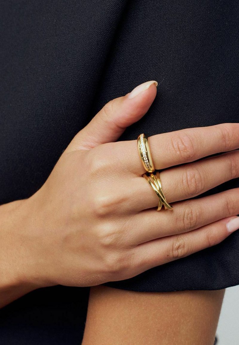 To goldringe på en hånd, den ene glat og rund, den anden med et snoet design. Hånden mod en mørk stofbaggrund.