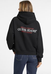 Čierna mikina s kapucňou a veľkým bielym a červeným logom "GUESS JEANS" na chrbte, vybavená stiahnuteľnou kapucňou a rebrovanými manžetami a lemom.