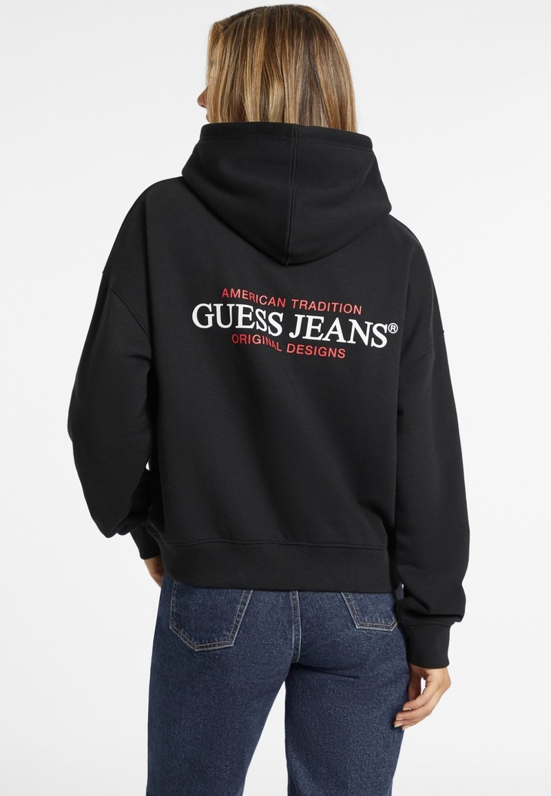 Čierna mikina s kapucňou a veľkým bielym a červeným logom "GUESS JEANS" na chrbte, vybavená stiahnuteľnou kapucňou a rebrovanými manžetami a lemom.