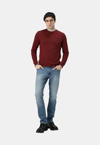 Maglione lungo rosso scuro a maniche lunghe con scollo rotondo, abbinato a jeans azzurri e scarpe nere. Il maglione presenta un piccolo logo.