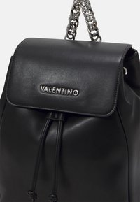 Svart läderryggsäck med ett vikbart lock, dragsko-stängning och ett silverfärgat kedjerem. Har varumärkesnamnet präglat på framsidan.