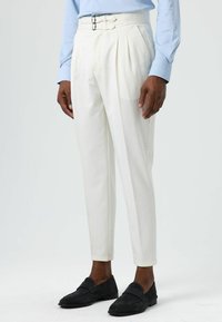 White pleated trousers with two metal buckles at the waist. Made from smooth fabric, featuring tapered legs and side pockets. Black loafers worn.
