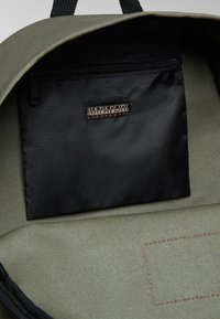 Zelená texturovaná podšívka batohu s černou zipovou kapsou s označením značky. Všimněte si kontrastního červeného prošívání na látce.