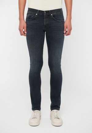 Mann in dunkelblauen Slim-Fit-Jeans und weißen Sneakern, der vor einem schlichten hellen Hintergrund steht.