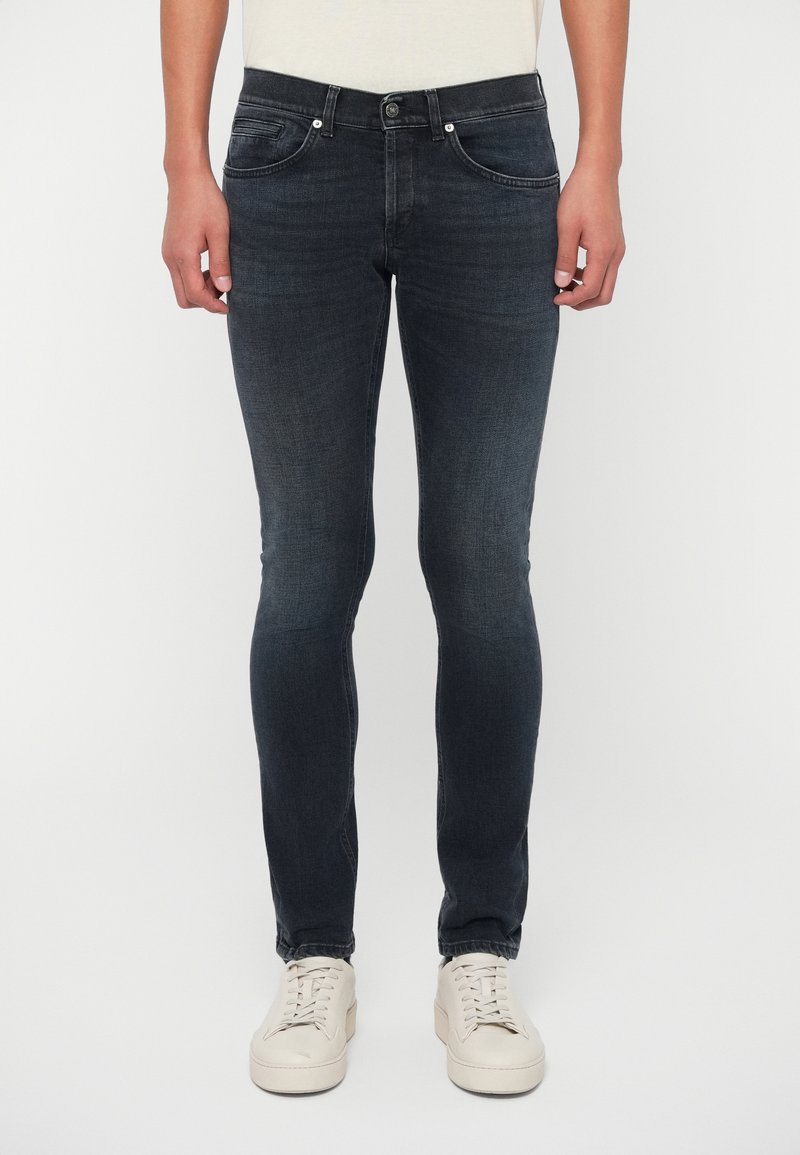 Άνδρας ντυμένος με σκούρα τζιν σε στενή γραμμή και λευκά αθλητικά παπούτσια, που στέκεται απέναντι σε μια φωτεινή απλή φόντο.