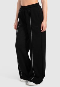 Sorte sweatpants med bred benvidde og en tekstureret overflade, der har hvid sidepiping og en elastisk talje. Logoet er trykt på forsiden.