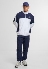 Sötétkék és fehér sportzakó, magas gallérral, cipzáras záródással és hozzáillő nadrággal. A cipőkön található fényvisszaverő részletek kontrasztot adnak. Hétköznapi sportos öltözet.