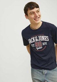 Tamnomodra bombažna majica s kratkimi rokavi, ki ima velik bel in rdeč grafični tisk z napisoma "JACK & JONES" in "90 DIV." Oblika z okroglim izrezom.