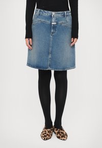 Personne portant une jupe en jean bleue, des collants noirs et des chaussons imprimé léopard, debout devant un fond blanc uni.