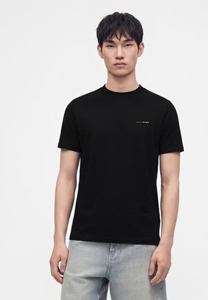 Ung mand iført en ensfarvet sort t-shirt med korte ærmer og lyseblå jeans, der står foran en hvid baggrund.