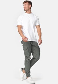Jeune homme marchant, portant un T-shirt blanc uni, un pantalon cargo gris avec des poches, et des baskets blanches avec des détails noirs.