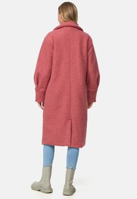 Manteau rose texturé à coupe décontractée, col oversize et manches longues. Porté avec un jean bleu clair et des bottines grises.