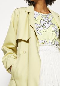 Lys gul trenchcoat med løs pasform og foldet krave, parret med en blomstermønstret top i pastelgul og hvid. Tekstureret hvid nederdel.