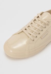 Beige patent skinnesko med rund tå, teksturert gummisåle og flate snører. Har en liten logo på siden nær snørene.