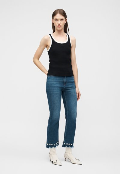 Γυναίκα που φοράει μαύρο-άσπρο αμάνικο τοπ, κοντά μπλε τζιν με διακόσμηση με λουλούδια και άσπρα μποτάκια με τακούνι, στέκεται μπροστά σε απλό φόντο.