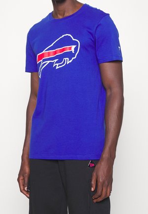 Homme portant un t-shirt bleu roi avec un logo stylisé de buffle blanc et rouge sur la poitrine, associé à un pantalon noir orné d'un écusson rose et noir.