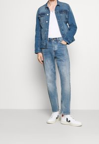 Jeansjacka i medelblå färg med knäppningar, bärs över en vit T-shirt, ihop med ljusblå smalpassande jeans och vita sneakers med svarta detaljer.