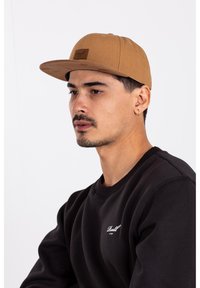 Brown cap with a flat brim and a rectangular logo patch, worn by a person in a black sweatshirt, featuring ribbed cuffs and neckline.