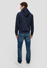 Sudadera azul marino con una gran capucha y puños acanalados, combinada con vaqueros azul desgastado y zapatillas blancas, vista desde la parte de atrás.
