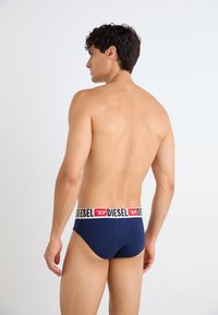 Dunkelblaue Unterwäsche mit einem weiße und roten Logo-Bund, glattes Material, körperbetontes Design, von hinten an einem männlichen Modell gezeigt.