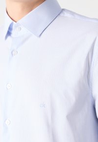 Camisa de botones a rayas azul claro hecha de algodón, con un cuello clásico y un logo bordado en el pecho.