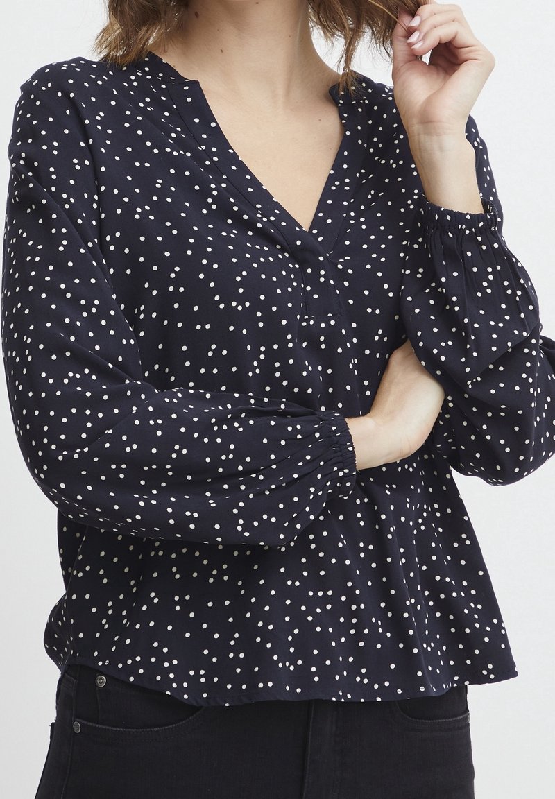 Femme portant un chemisier bleu marine à pois blancs et manches longues, associé à un pantalon noir, sur un fond uni.
