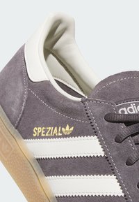 Zbliżenie na szary zamszowy but sportowy Adidas Spezial z białymi paskami, kremową podszewką, czarnymi sznurowadłami i gumową podeszwą.