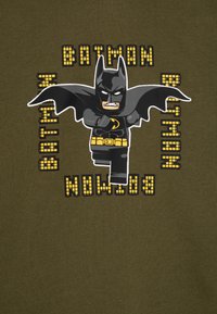 Žalia medžiaga su animacinio Batman personažu juodos ir geltonos spalvų, šoninėse esančiomis tekstu "BATMAN" pikseliu fonte.