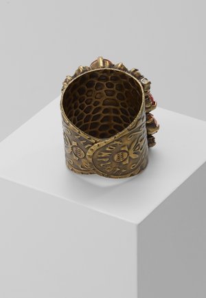 Anillo ancho de latón con un intrincado patrón grabado y un interior texturizado, colocado sobre un pedestal blanco frente a un fondo liso.