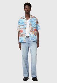 Kurzärmliges Hemd in Hellblau mit Wellen- und Vogelmustern, kombiniert mit hellblauen Jeans und schwarzen Schuhen. Glatte Textur.